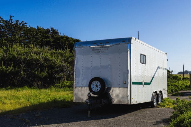 A white enclosed trailer parked on gravel beside a grassy hillside